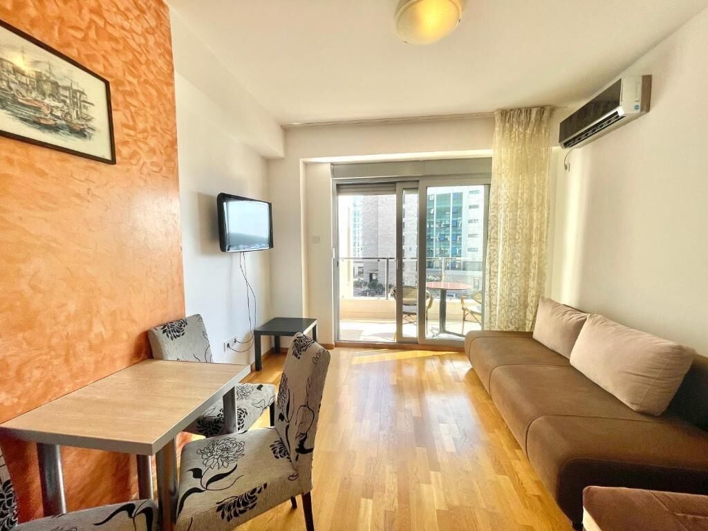 Een woonkamer met een bank, een eettafel met stoelen, een flatscreen televisie en een groot raam met uitzicht op de stad.