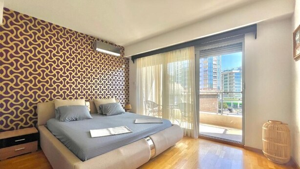 A modern bedroom with a large bed, a patterned wall, and a view of the city through a sliding glass door. - Comfort Apartments Aleksic (Budva)