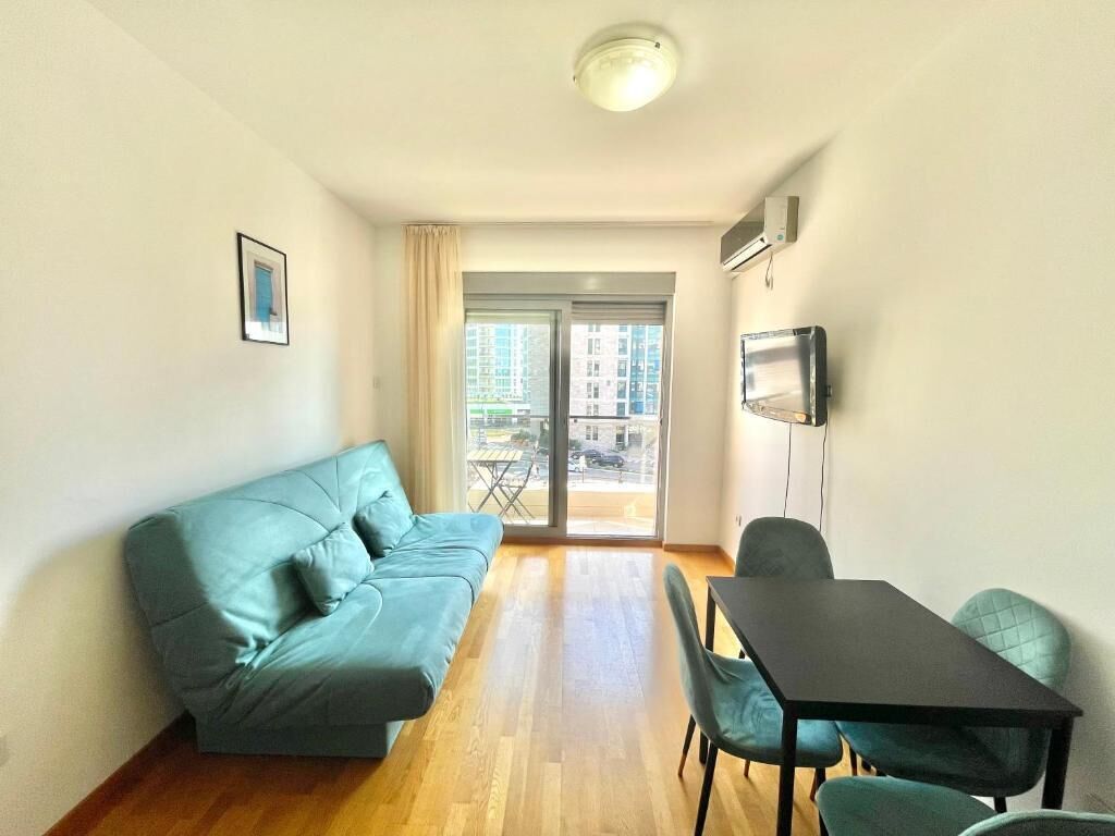 Un salon moderne avec un canapé, une table à manger et des chaises. Une grande fenêtre offre une vue sur les bâtiments environnants.
