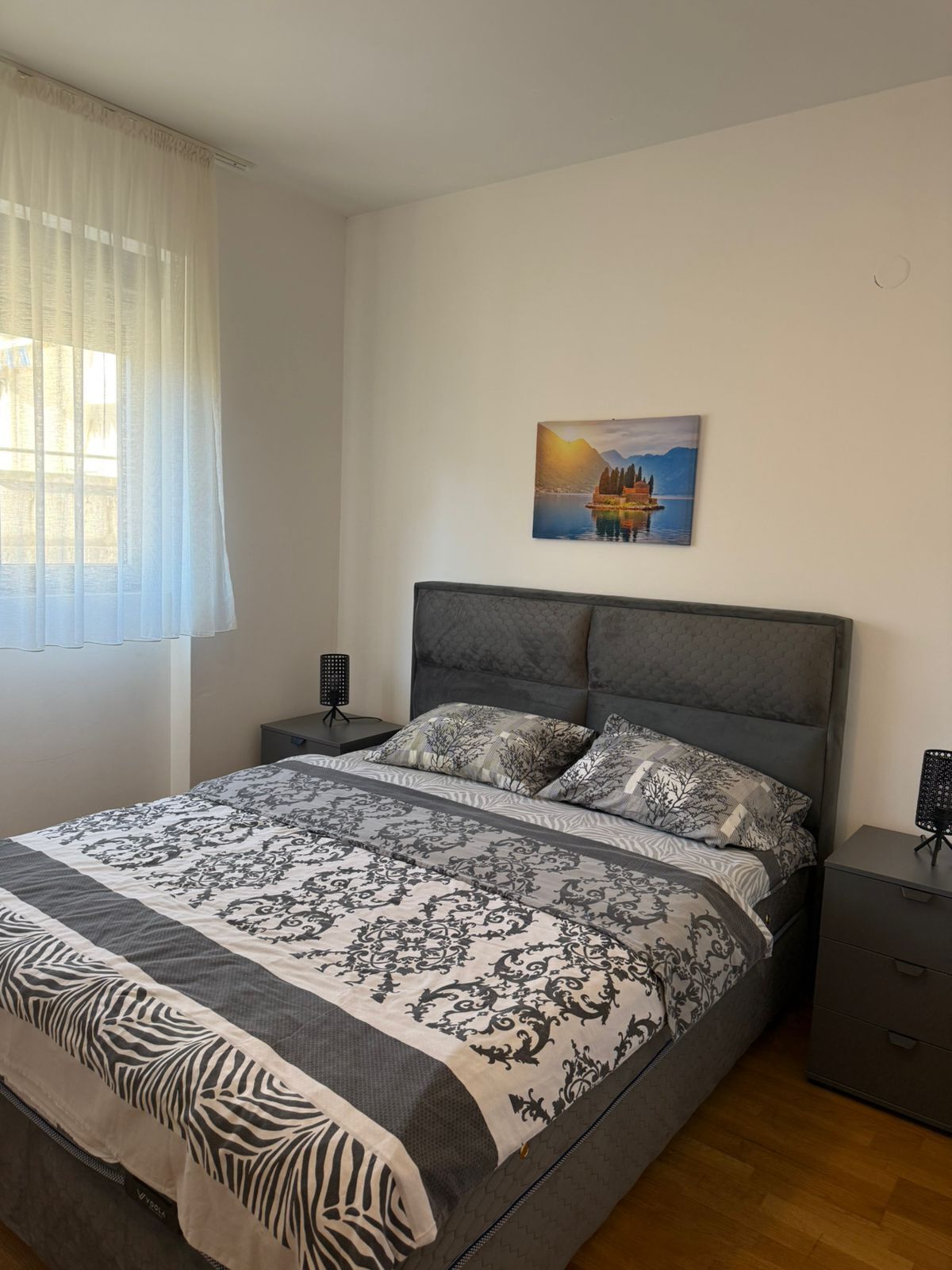 Un dormitorio con cama, mesita de noche, cortinas en la ventana y un cuadro colgado en la pared.