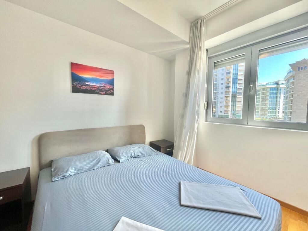 A bedroom with a bed, a nightstand, a window with curtains, and a cityscape painting on the wall.