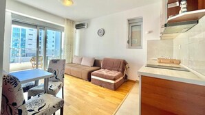 A modern living room with a sofa, a dining table with chairs, a small kitchen area, and a large window offering a view of the city. - Comfort Apartments Aleksic (Budva)
