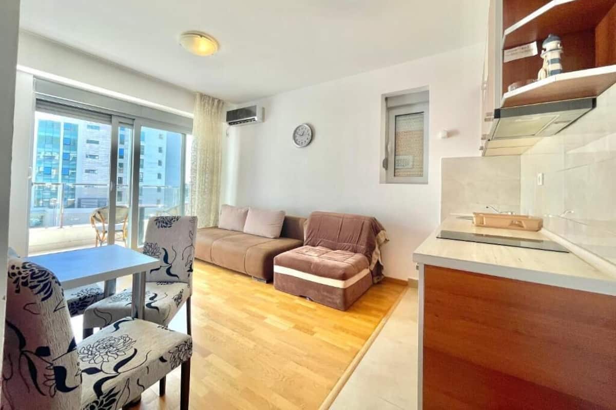 A modern living room with a sofa, a dining table with chairs, a small kitchen area, and a large window offering a view of the city.