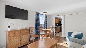 A modern living room with a wooden coffee table, a sofa, and a dining area with a round table and chairs. A flat-screen TV is mounted on the wall, and there is a window with patterned curtains. - Seaside King Suite 12th Flr Free Breakfast (Myrtle Beach)