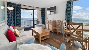 A modern living room with a sofa, coffee table, and wooden chairs. There is a balcony with a view of the ocean. - Seaside King Suite 12th Flr Free Breakfast (Myrtle Beach)