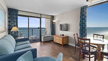 A room with a balcony, a sofa, a dining table with chairs, a TV, and a dresser.