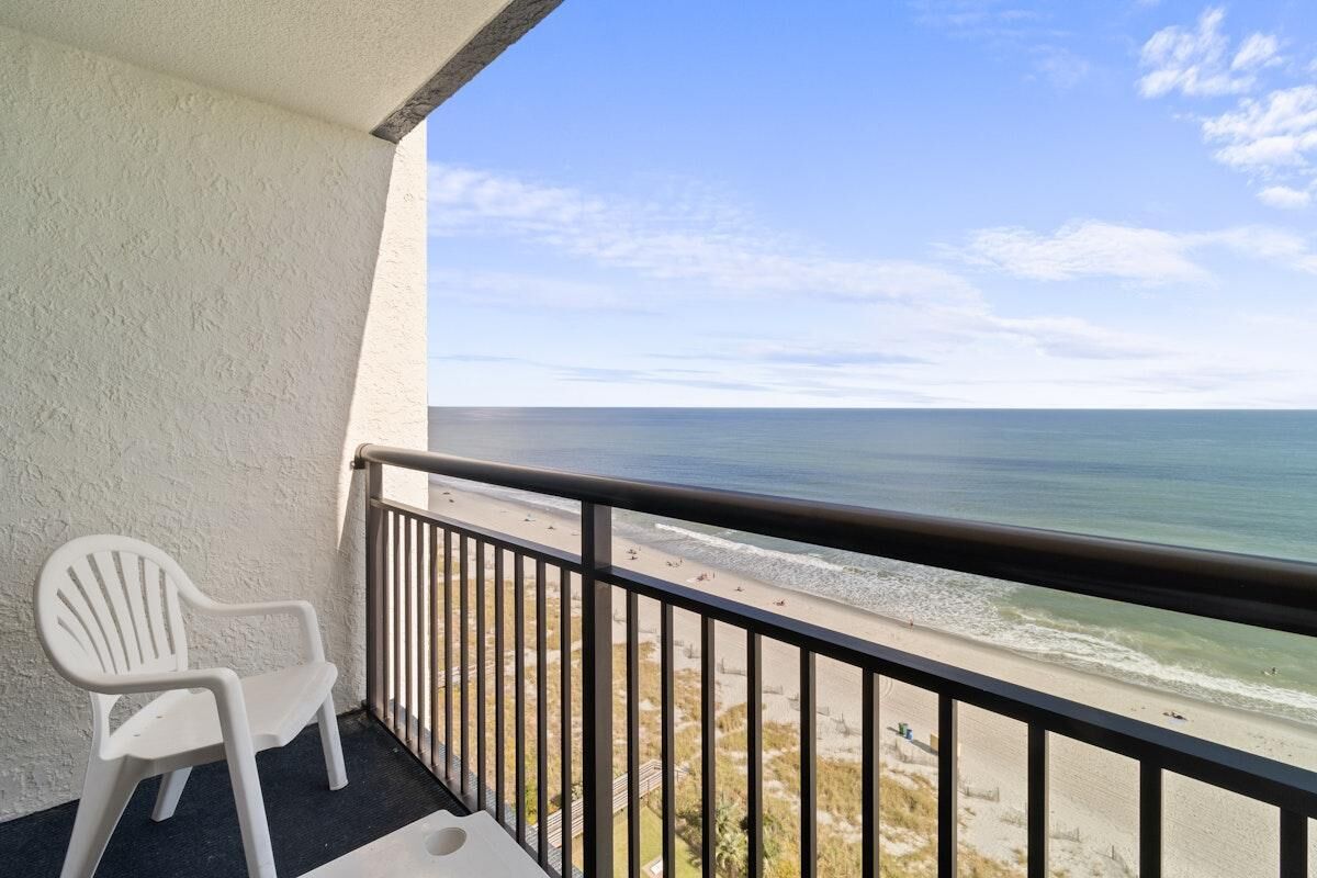 Un balcón con una silla de plástico blanco, barandilla y vista a la playa y el océano.