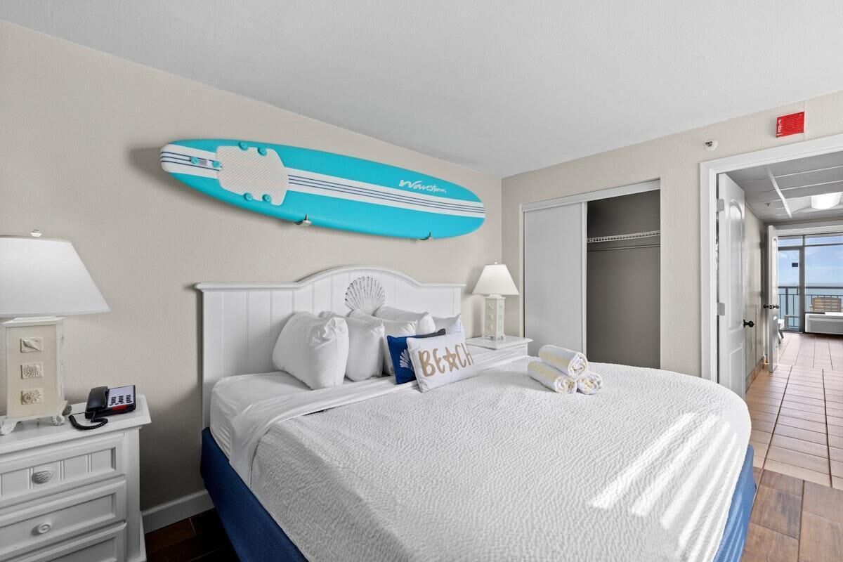 Ein Hotelzimmer mit Bett, Nachttisch, Lampe, Telefon, einer Surfbrett-Dekoration und Blick auf den Balkon.