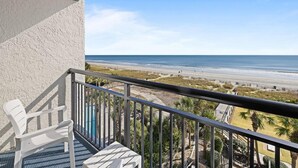 A balcony with a view of the beach, a white chair, and a small table.
