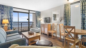 Ein modernes Wohnzimmer mit einer Couch, einem Esstisch und Stühlen. An der Wand ist ein Fernseher angebracht, ein kleines Schränkchen steht dort und ein Fenster mit Vorhängen ist zu sehen.