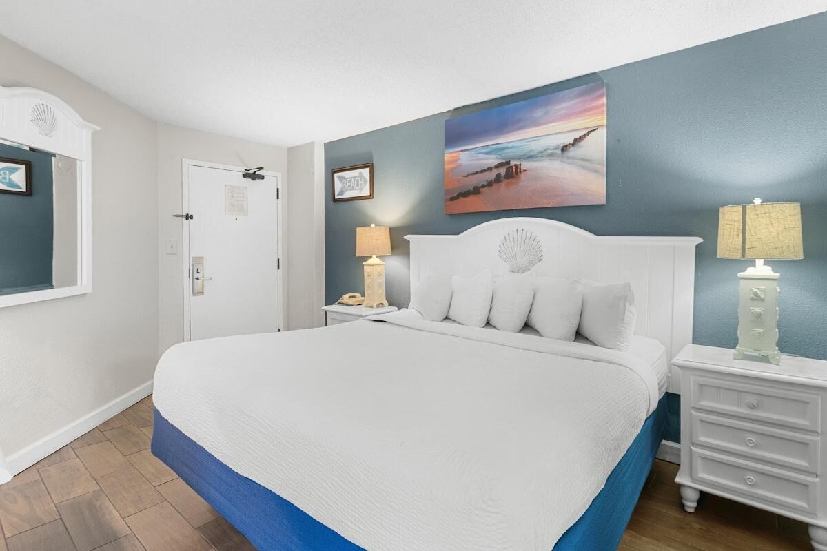 A bedroom with a large bed, a nightstand with a lamp, a wall-mounted artwork, and a door with a sign.