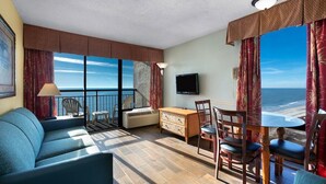 A room with a balcony, a sofa, a table, chairs, a TV, and a view of the ocean.
