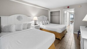 Et hotelværelse med to senge, et natbord med en lampe, trægulv og udsigt til gangen.