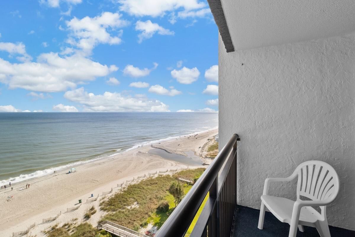 A balcony with a white plastic chair, overlooking a beach with a fence and a boardwalk.
