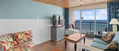 Una habitación con techo de madera, un sofá estampado con motivos florales, una mesa, un armario, un televisor y vistas al mar a través de amplios ventanales.