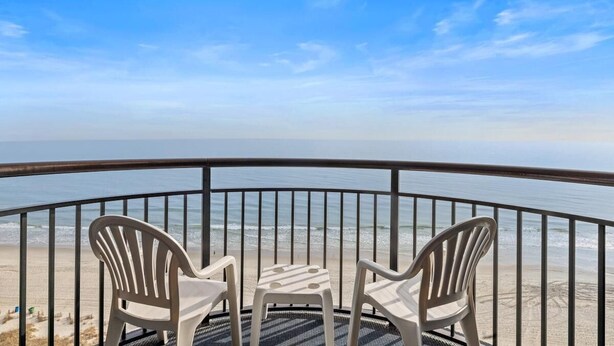 A balcony with two white plastic chairs and a small table, overlooking a beach with a clear sky. - Airy Oceanfront Suite with Breakfast 14th Floor (Myrtle Beach)