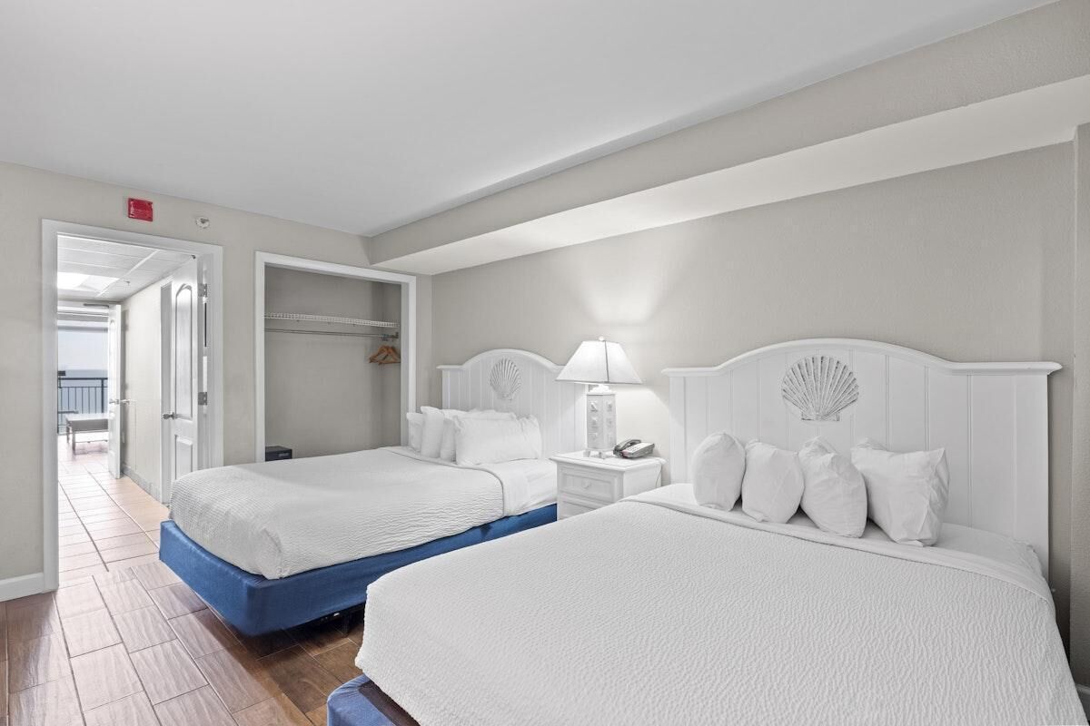 Habitación de hotel con dos camas, un vestidor, una mesita de noche con lámpara y vistas al pasillo.