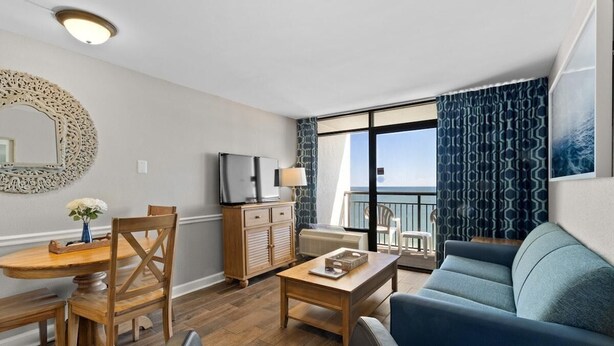 A modern living room with a wooden dining table, a sofa, a TV, and a view of the ocean through large windows. - Beachy Oceanfront Ste with Breakfast 14th Flr (Myrtle Beach)