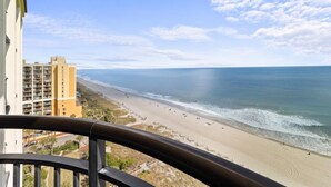 A beach view from a balcony with a clear sky, a sandy beach, and a building in the background.