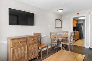 Un comedor con mesa y sillas de madera, un mueble de madera, un televisor de pantalla plana montado en la pared y una cocina visible a través de un vano abierto.