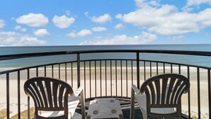 A balcony with two Adirondack chairs and a small table, overlooking a beach with a clear sky.