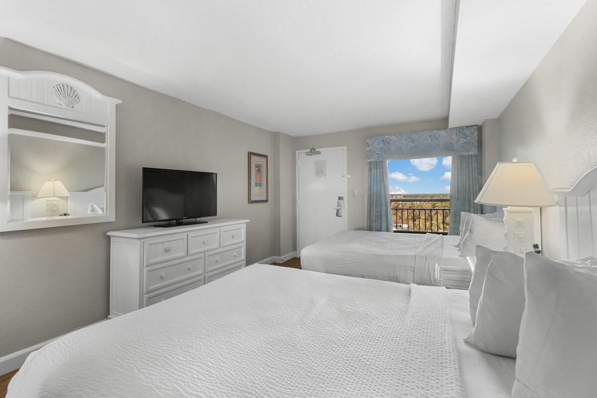 A hotel room with two beds, a TV, a dresser, and a balcony with a view.