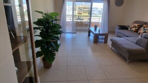 Un salon spacieux avec un canapé gris, une table basse et une grande plante verte. On aperçoit, à travers la fenêtre, un balcon avec une table et des chaises.