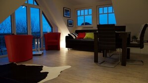 Een moderne woonkamer met een zwarte leren bank, rode stoelen, een eettafel en een kleden van koeienhuid. Grote ramen bieden uitzicht op het omliggende landschap.
