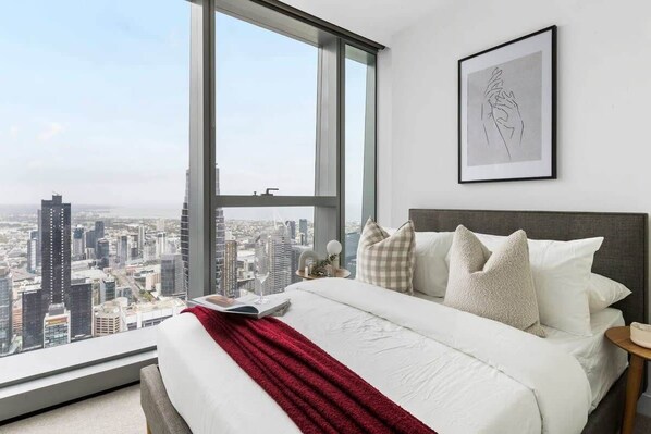 A modern hotel room with a large window offering a city view, a neatly made bed with white linens and a red throw, and a framed artwork on the wall.