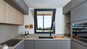 A modern kitchen with a built-in oven, a window with a view, and a countertop with wine bottles and glasses. - Panoramic luxury villa with private pool in Sampieri - Di Casa In Sicilia (Sampieri)