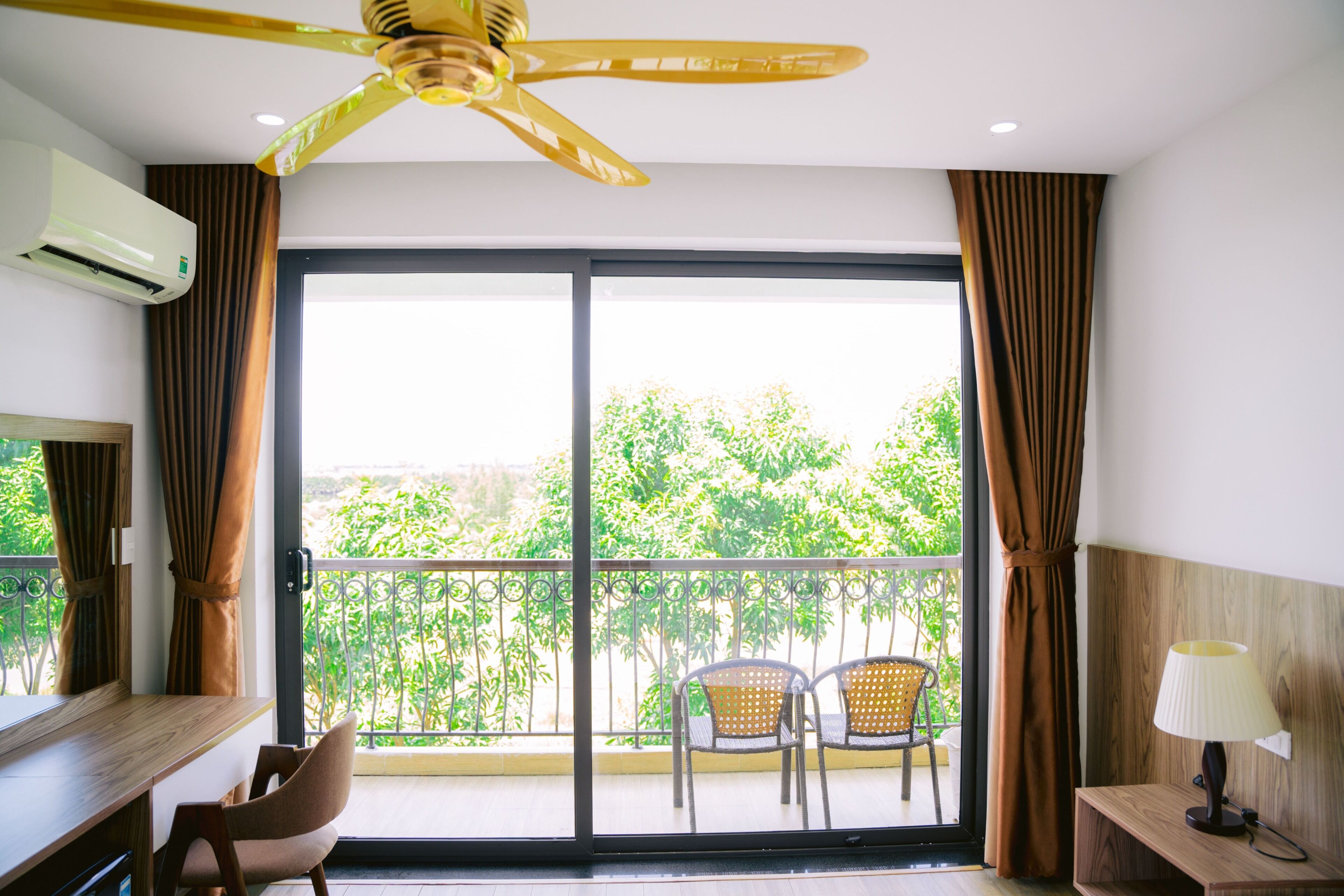 Habitación de hotel con ventilador de techo, balcón con sillas y vistas a zonas verdes.