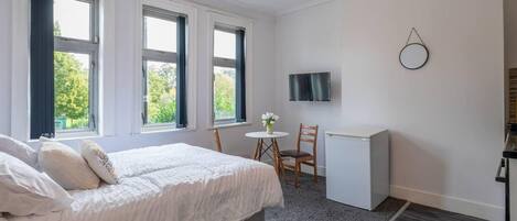 A bedroom with a bed, a desk, a chair, a TV, and large windows.