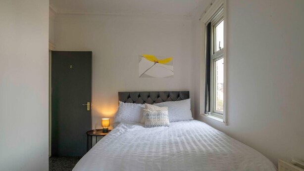 A bedroom with a bed, a nightstand, a lamp, a window, and a door.