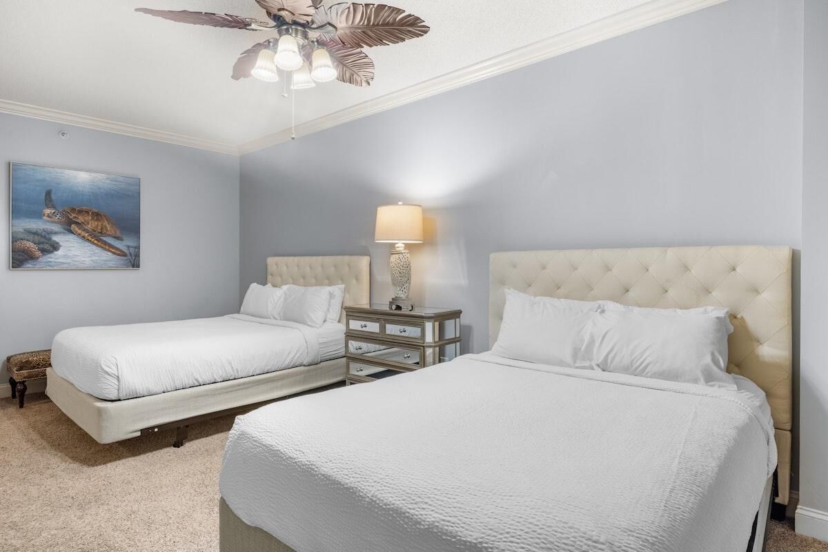 A hotel room with two beds, a ceiling fan, a nightstand with drawers, a lamp, and a painting of a sea turtle.