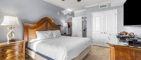 A bedroom with a bed, a nightstand, a lamp, a ceiling fan, a TV, and a door.