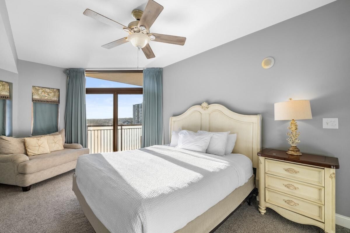 Habitación de hotel con cama, mesita de noche, ventilador de techo, sofá y vista a la ciudad.