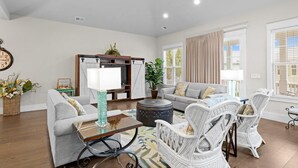 A spacious living room with a grey sofa, a coffee table, wicker chairs, a side table, a potted plant, and a large window with curtains. - Cozy 4 Bedroom Beach House Close to the Ocean (North Myrtle Beach)