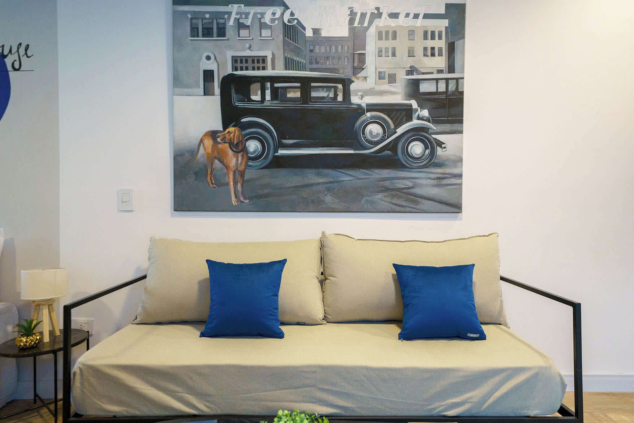 Une pièce avec un canapé beige, deux coussins bleus, une petite table avec une lampe et un tableau mural représentant une voiture ancienne et un chien.