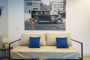 A room with a beige sofa, two blue pillows, a small table with a lamp, and a wall painting of a vintage car and dog.