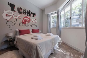 Un lit bien fait, avec du linge de lit blanc et des coussins rouges, un mur orné d’un texte motivant, une petite table avec une lampe et une grande fenêtre avec des rideaux.
