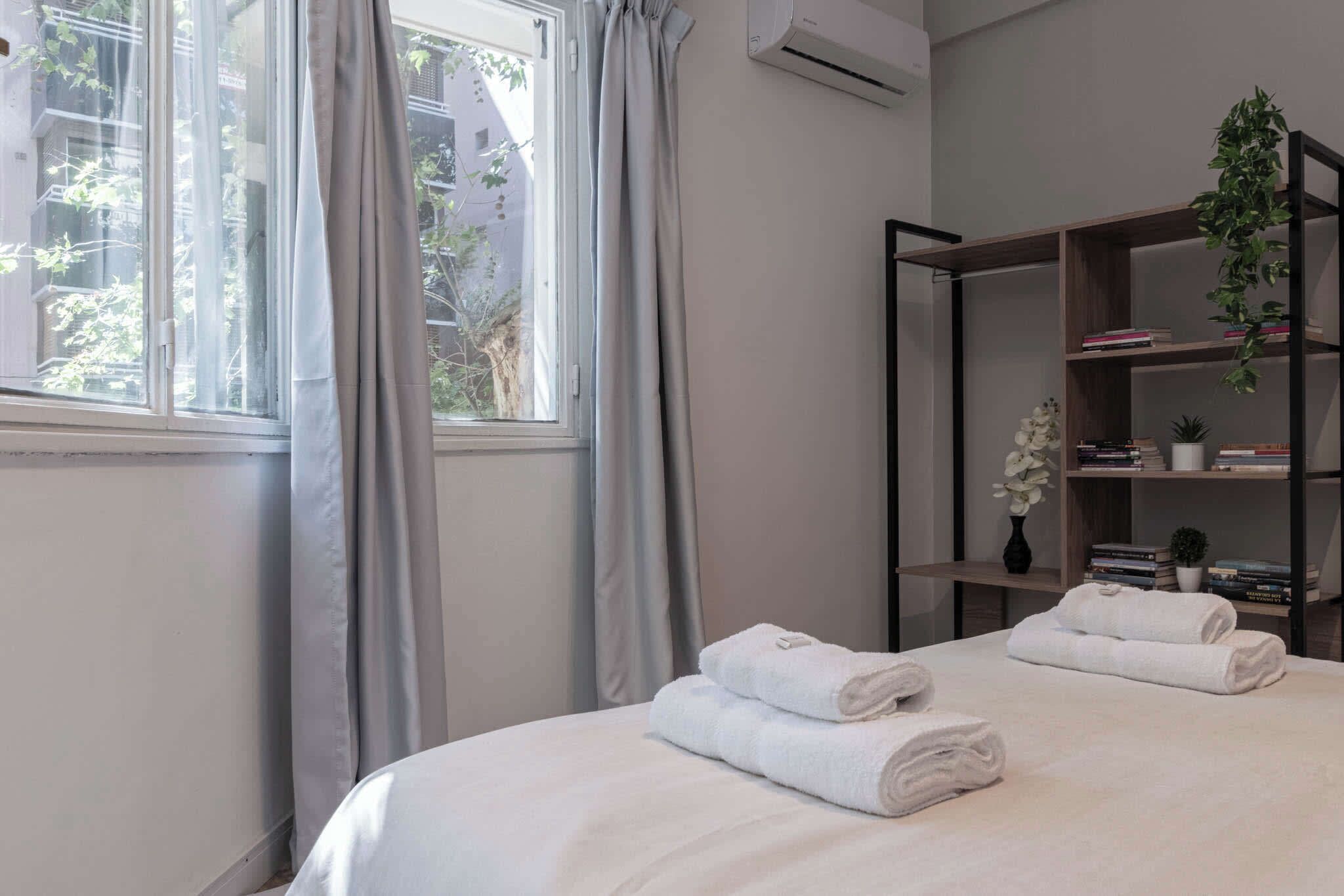A hotel room with a bed, two rolled towels, a bookshelf with books and plants, and a window with curtains.