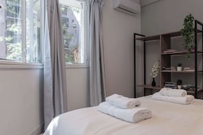 Habitación de hotel con cama, dos toallas enrolladas, una estantería con libros y plantas, y una ventana con cortinas.
