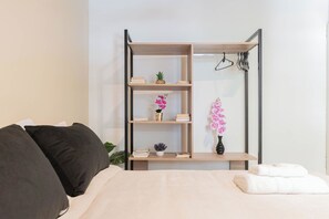 Et soveværelse med en seng, et natbord, en reol med bøger og en lille plante, samt en vægmonteret bøjle med tøj.