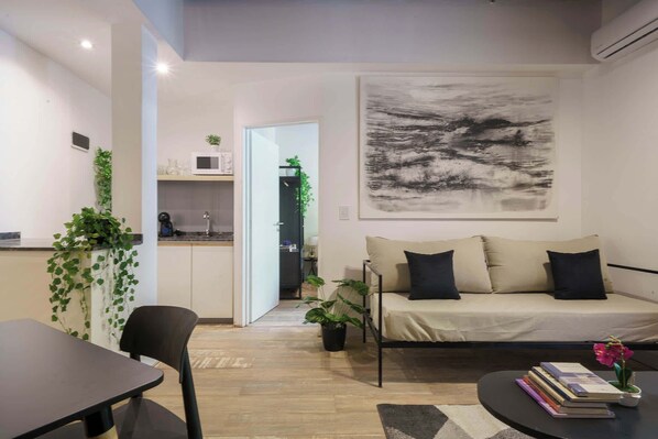 Una sala moderna con un sofá, una mesa de comedor y una mesa de centro. Se observa un área de cocina al fondo, con un microondas y una planta.