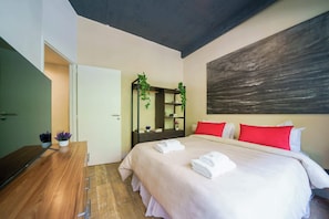 Une chambre à coucher moderne avec un grand lit, une commode en bois, un miroir et un ensemble d’oreillers rouge et blanc.