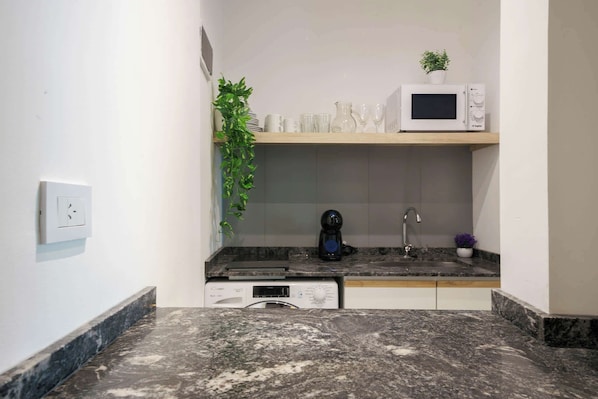 A compact kitchen with a microwave, coffee maker, and a small shelf with glassware.