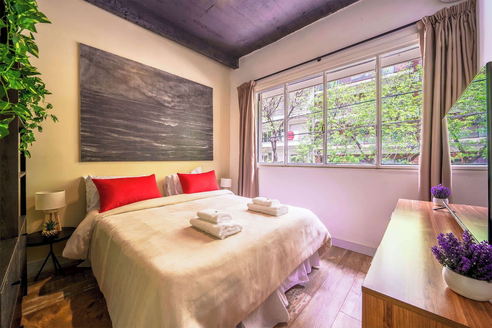 A hotel room with a bed, a large abstract painting, a window with curtains, a wooden desk, and a potted plant.