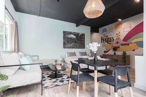 Una sala de estar moderna con sofá, mesa de comedor y sillas. Hay un mural en la pared y una ventana con cortinas.