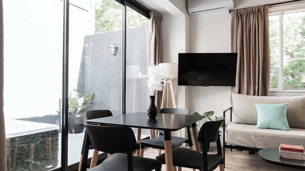 Un salon moderne comprenant un canapé, un téléviseur fixé au mur, une table à manger avec des chaises et une grande fenêtre avec des rideaux.