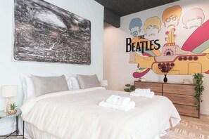 A bedroom with a bed, a nightstand, a lamp, a wall art of The Beatles, and a dresser with a plant.